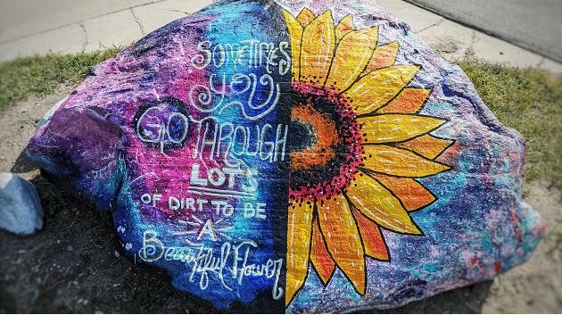 An art project undertaken by participants in Vista Maria's Journey to Success program. Large painted boulder, right half is half a sunflower the left half is blues and purples with "sometimes you go through lots of dirt to be a beautiful flower"