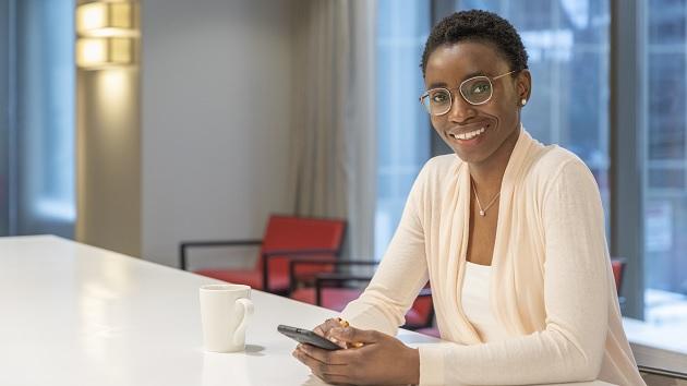 person sitting at a large table with phone in both hands and coffee mug next to them, smiling at the camera