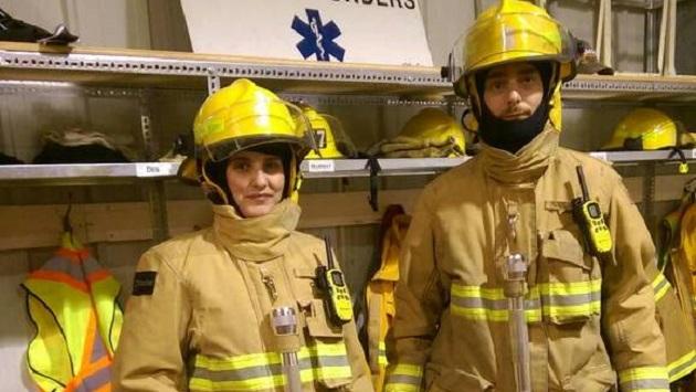 Two people in firefighter uniforms stand in front of a closet of safety equipment