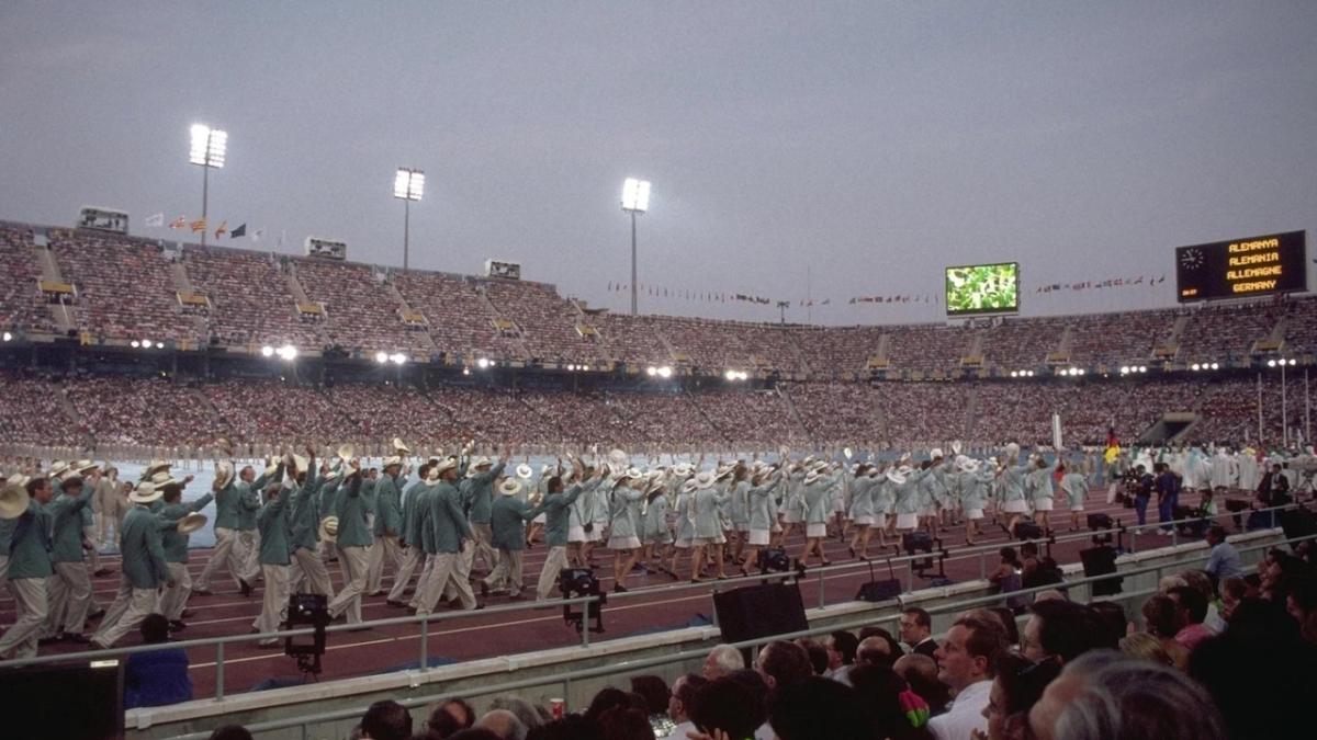 athletes dressed in matching uniforms walking along a track of a stadium. Stands filled with spectators.