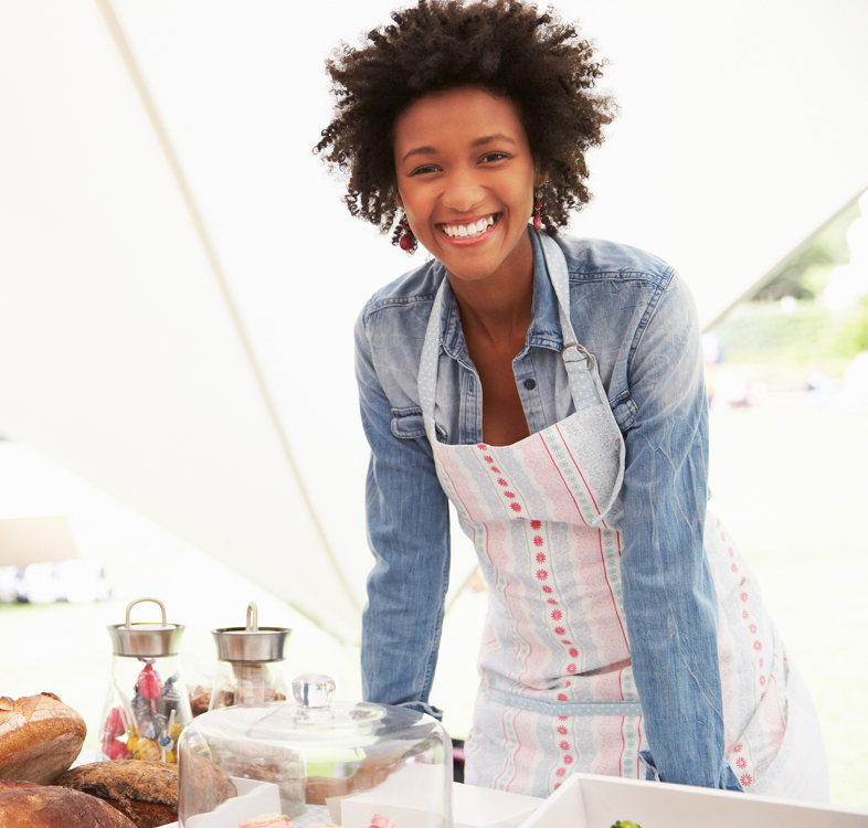 a person wearing an apron in a white tent, baked goods in front of them