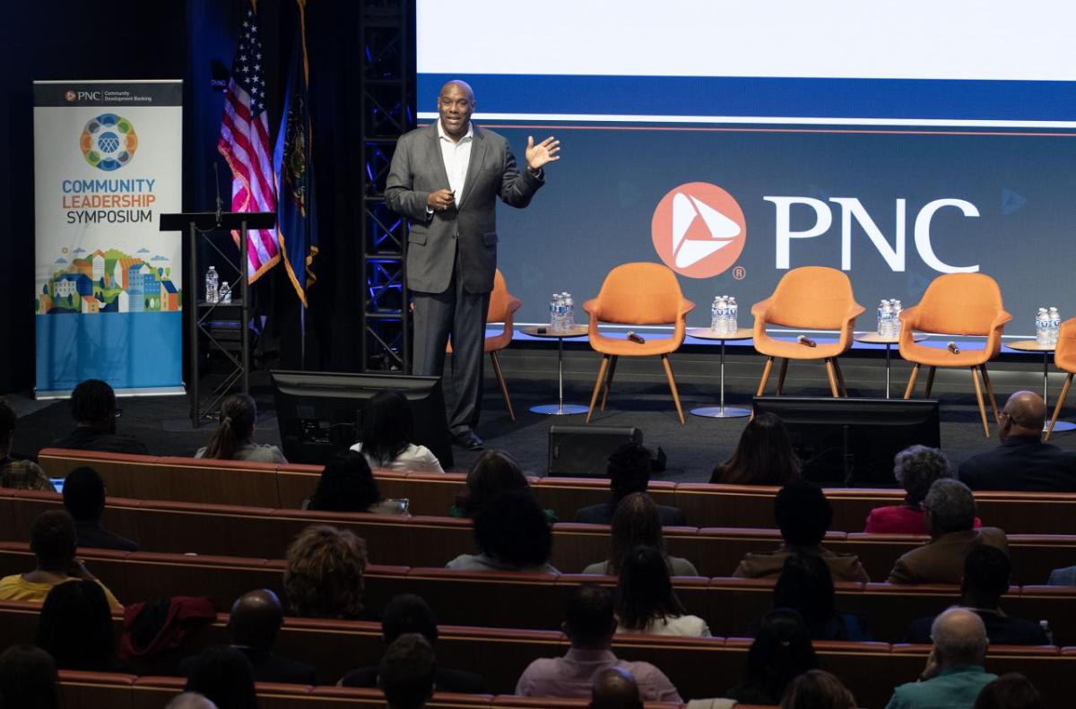 Richard Bynum standing on a stage in an auditorium of seated people. Chairs and PNC banner behind them.