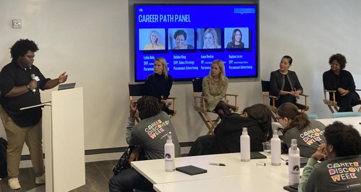 Four people seated with a display "Career path panel" behind them, all listening to a speaker at a podium to the left. Students seated at tables in the room.