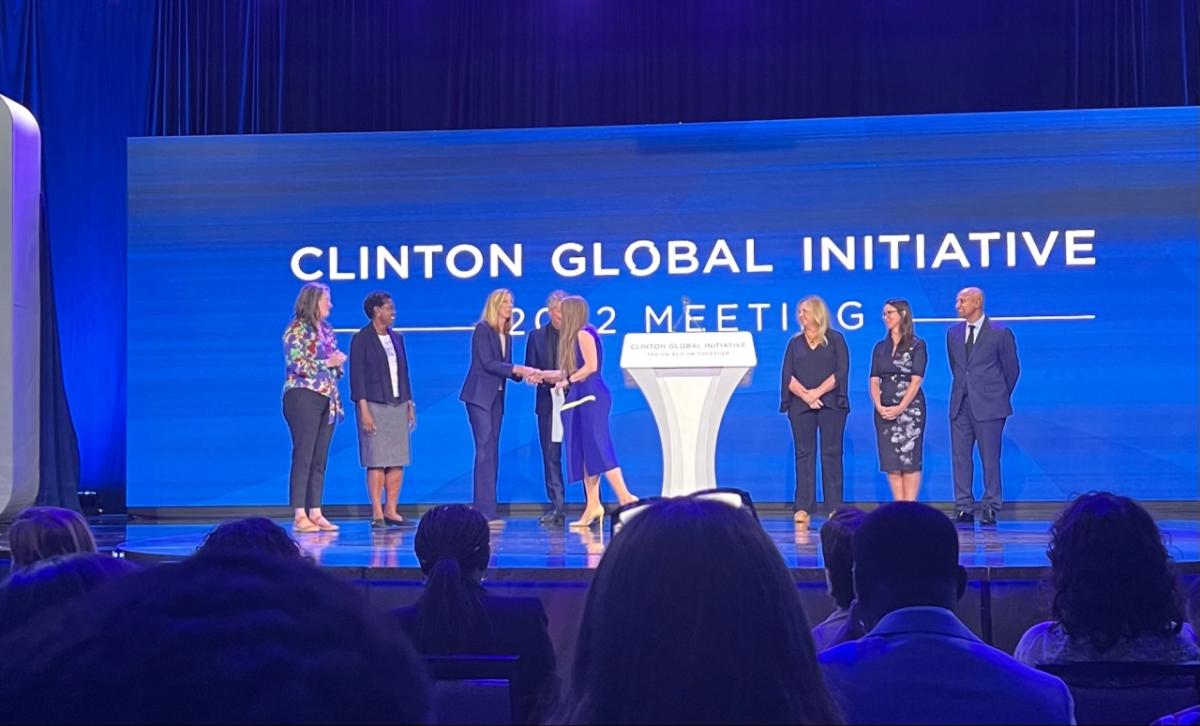Americares President and CEO Christine Squires shakes hands with Chelsea Clinton on stage at the Clinton Global Initiative 2022 Meeting in New York City. Photo by Yael Gottlieb for Americares. Image of eight people on stage in front of a backdrop that says, "Clinton Global Initiative"