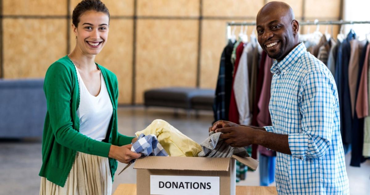 Two people holding clothing items in a "Donations" box. A rack of clothing hanging behind them.