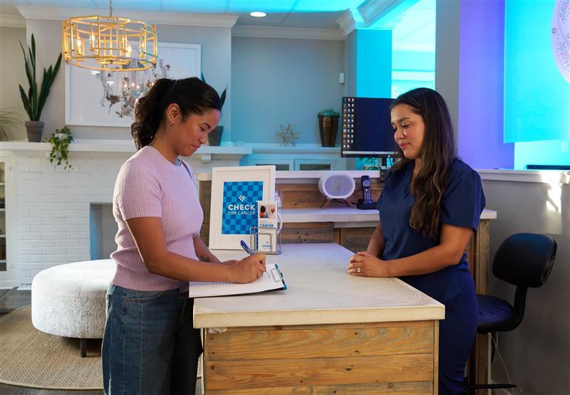 A person filling out a form at a clinic with another person behind a desk.