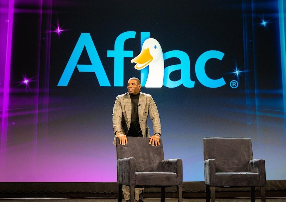 Coach Prime shown on stage with the Aflac logo.