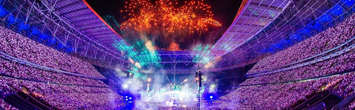 Image via Robert Mallows A huge stadium filled with spectators, a stage central is brightly lit, and orange fireworks fill the sky.