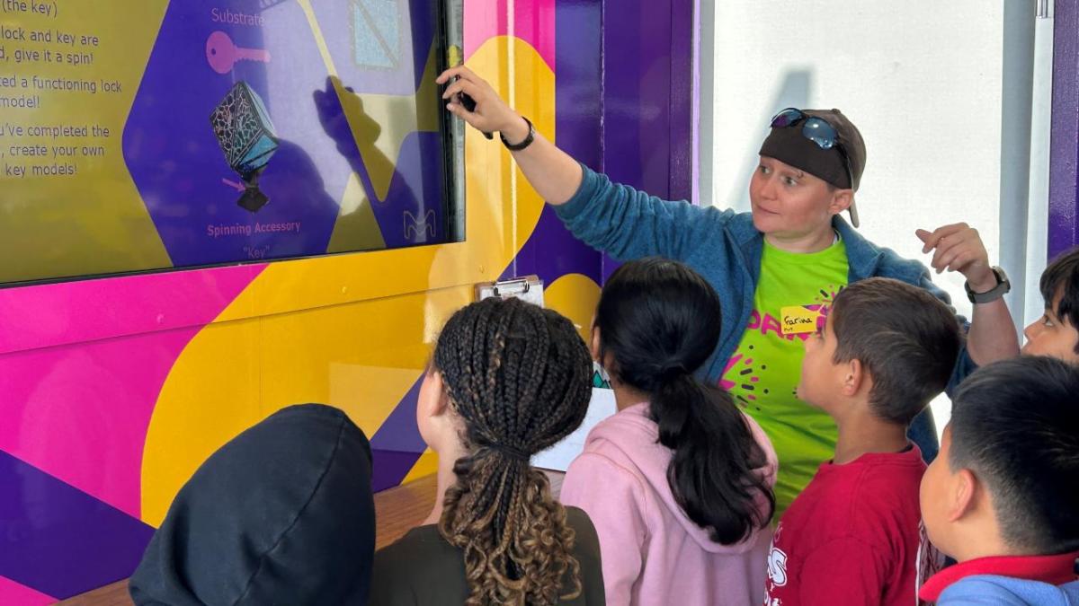 Volunteer inside the Curiosity Cube™ guiding a group of students through an interactive science activity on a TV screen.