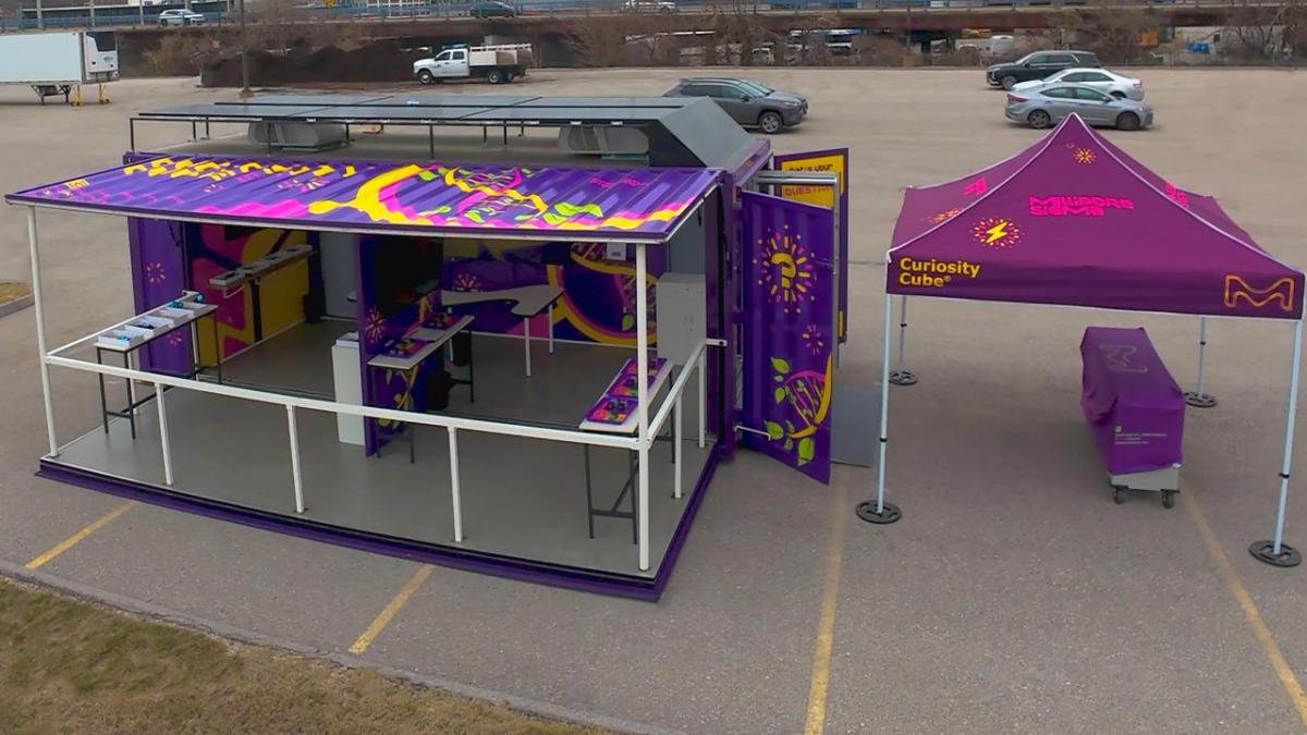 Aerial view of the Curiosity Cube™ mobile science lab with side panels open, revealing interactive experiment stations beneath a colorful canopy.