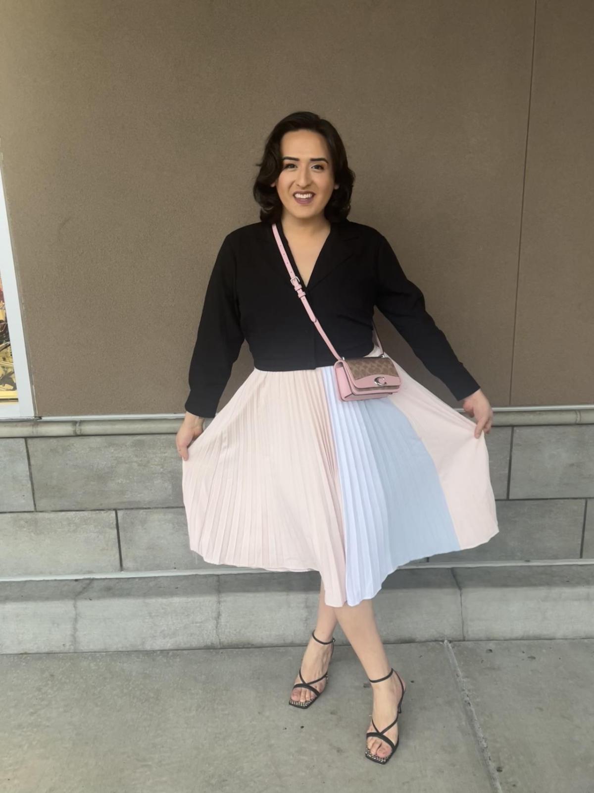 Curtlynn shows off her skirt, pleated pastel striped, strapy black heels and black v-neck top. She stands, smiling, in front of an exterior beige wall.