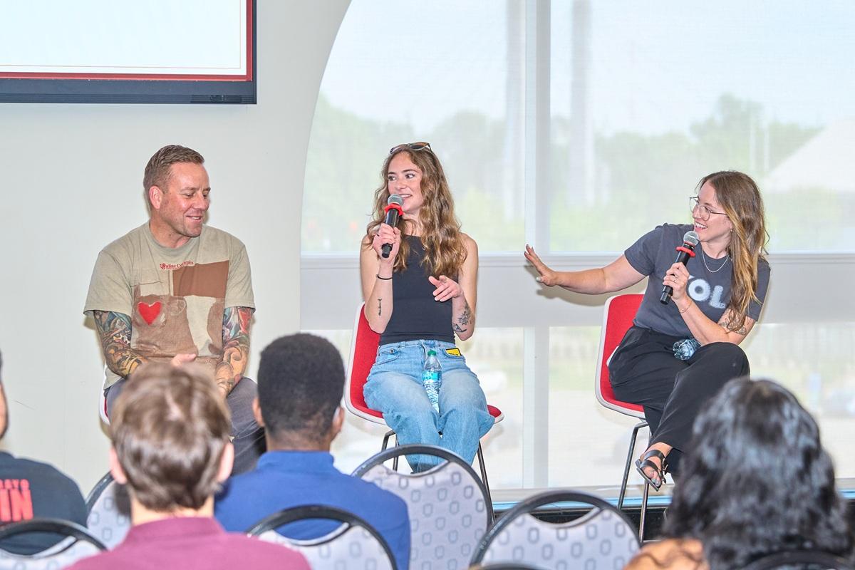 Three speakers sit onstage during a panel discussion, holding microphones and speaking to an audience seated in a conference room.