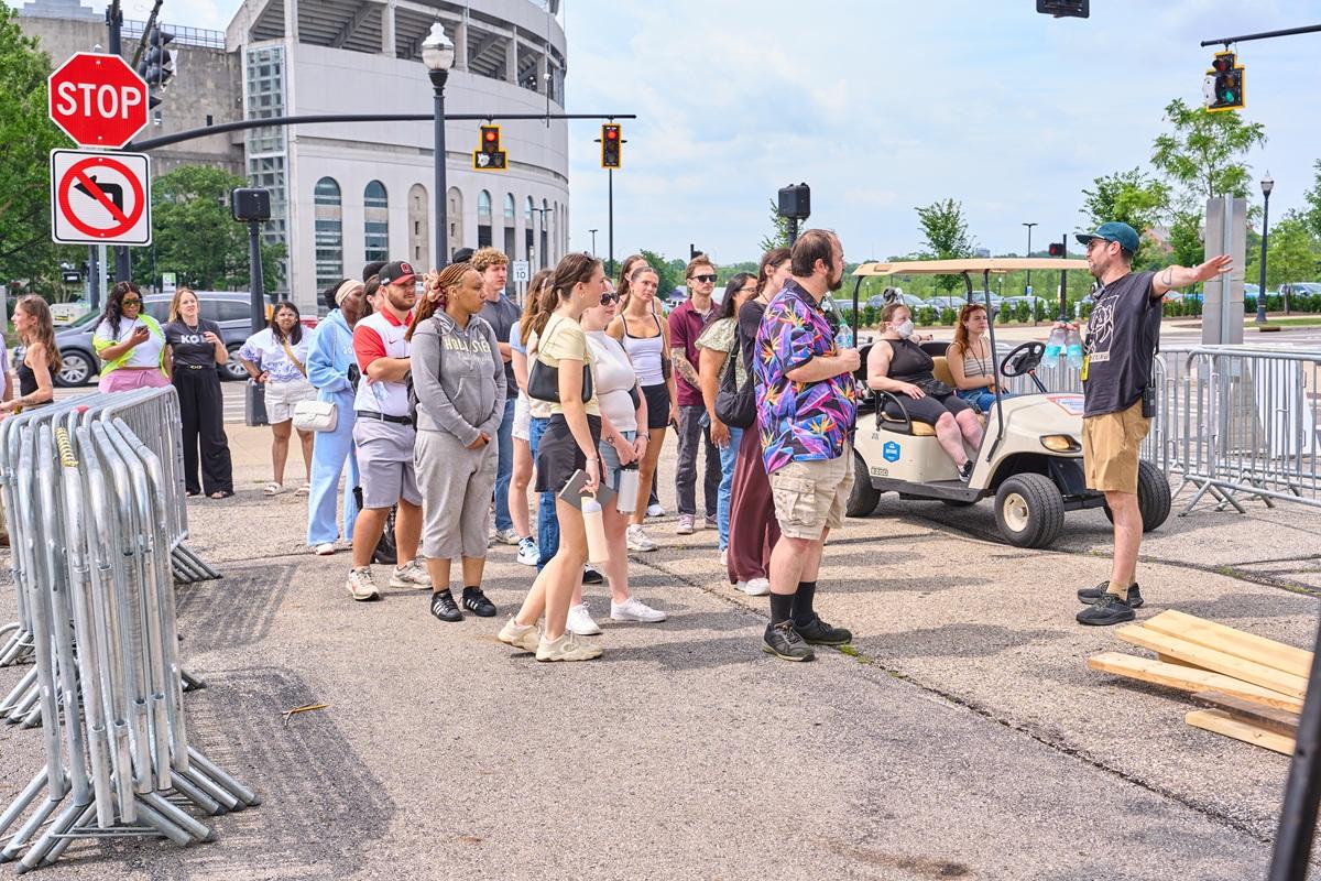 College program participants gather outdoors near crowd-control barricades as a staff member provides a venue operations briefing outside a stadium before an event.