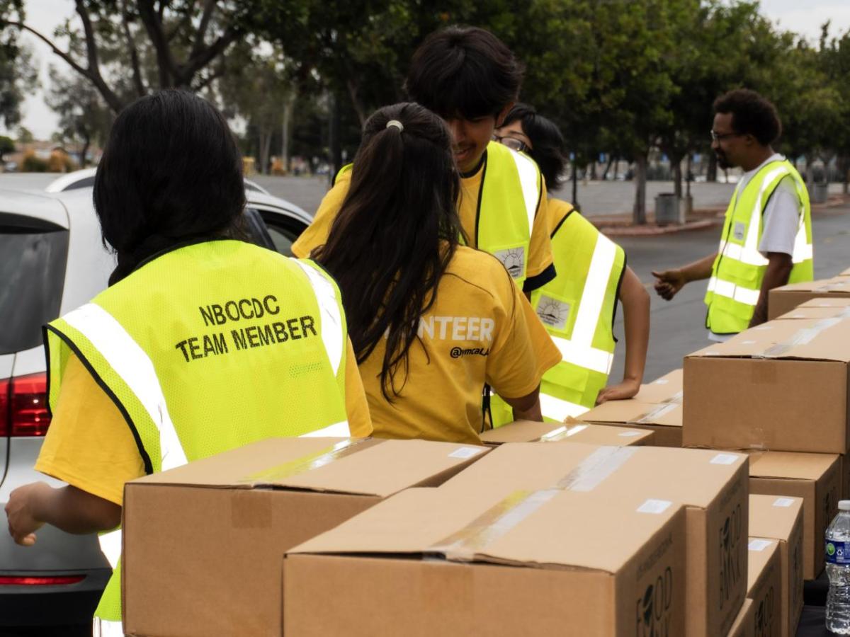 Volunteers in yellow vests gather around a long table stacked with sealed brown food bank boxes.