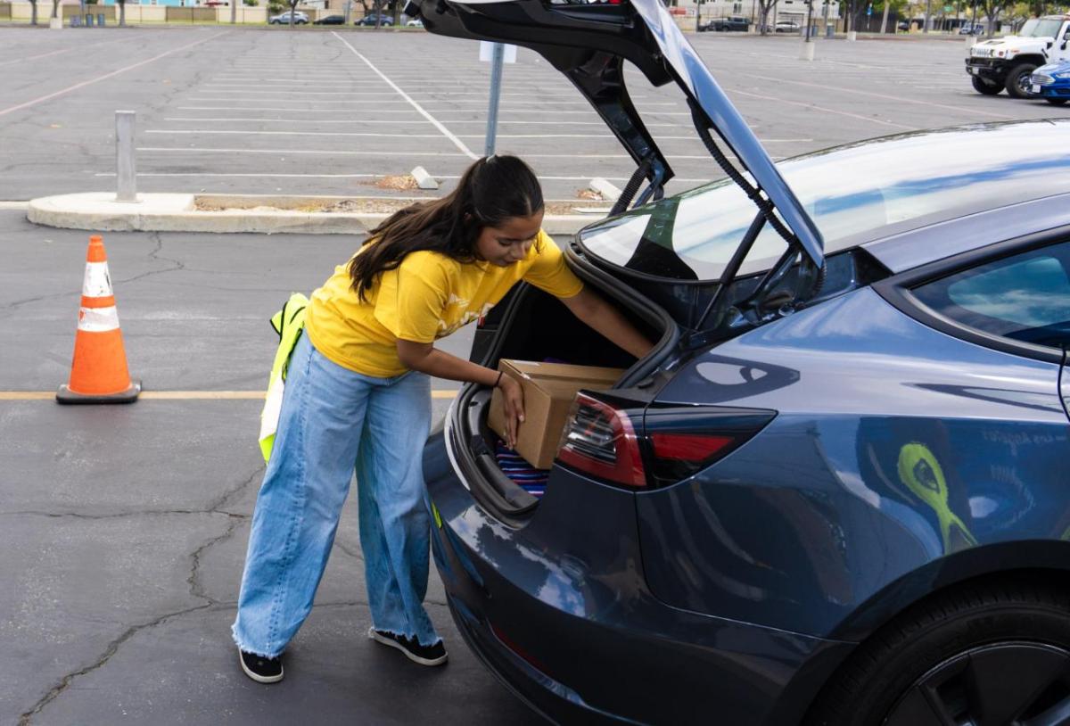 A volunteer in a yellow shirt places a brown box in the trunk of a blue car in a parking lot.