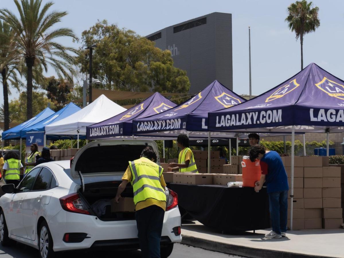 A food distribution site with LA Galaxy-branded canopies and volunteers loading boxes into a white sedan.
