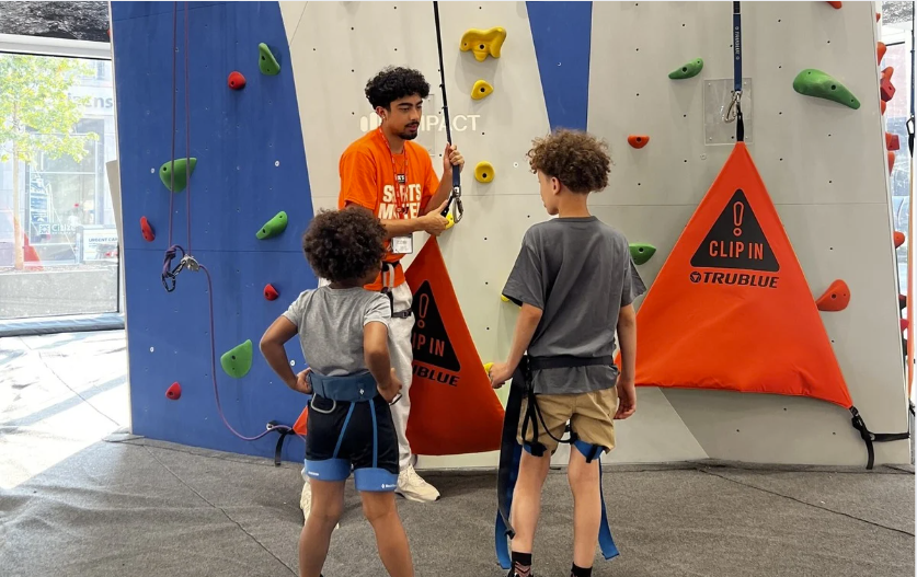 Two children about to be strapped in for a climbing wall adventure.