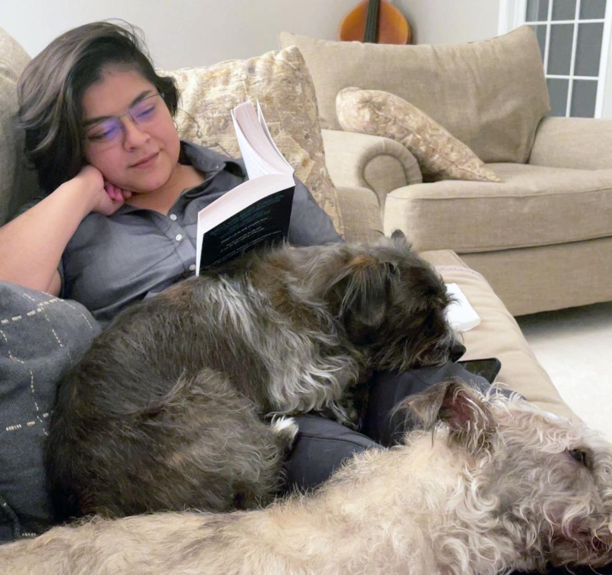 Denisse De la Cruz reading on a couch with two dogs curled up next to her