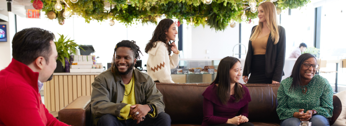 Group of people sitting inside on a couch or standing behind it, talking with each other. A cash register and bakery case behind them.