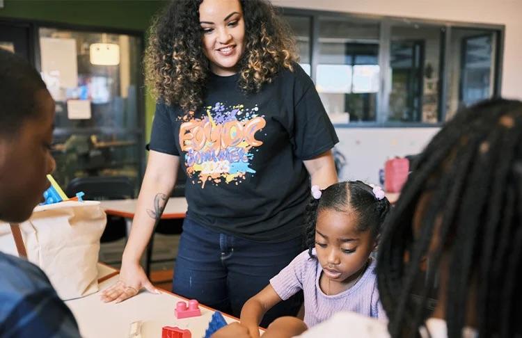 Person stood looking over a child who is playing with plastic bricks at a table