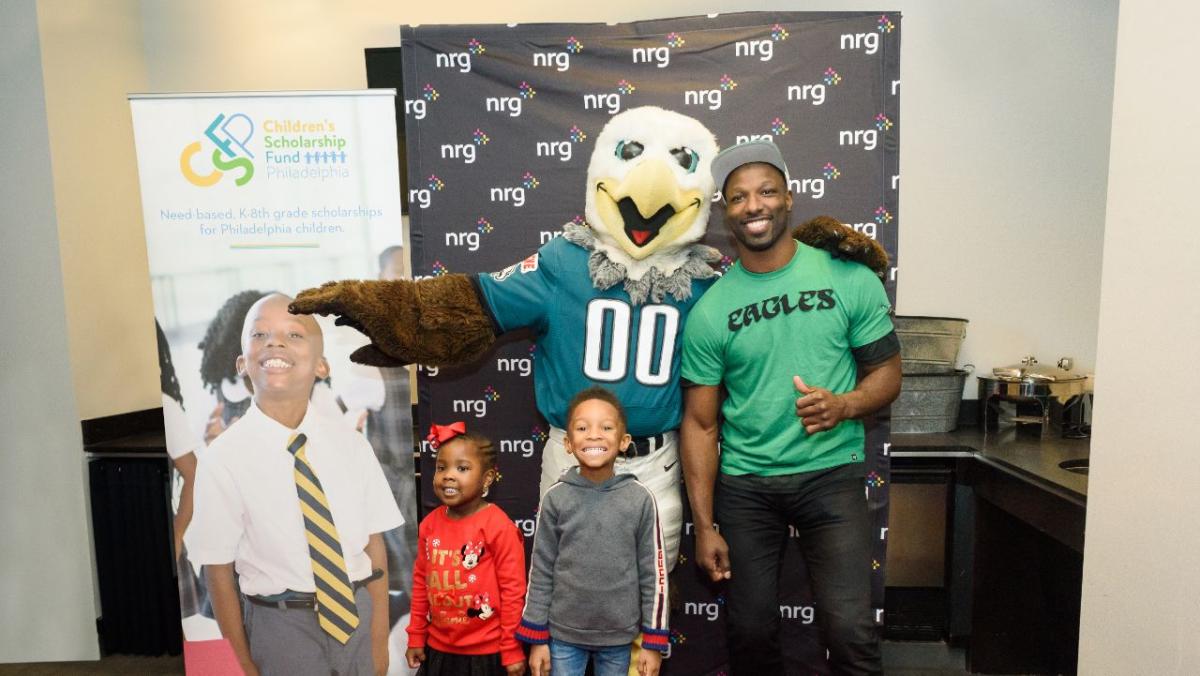 An adult, two children and Eagle mascot pose in front of an NRG screen and tall banner for CSFP.