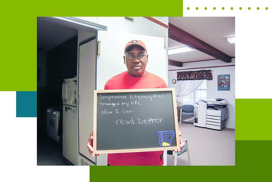 Person stands in a home setting holding a sign that reads "Improving literacy has changed my life. Now I can read better"