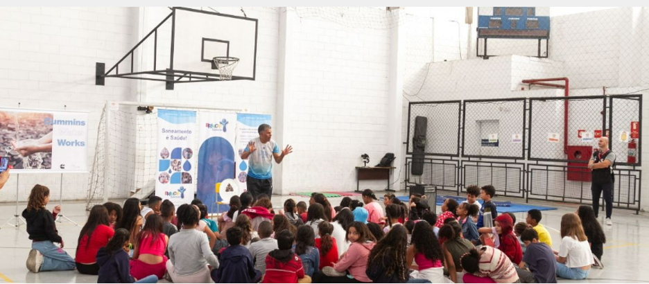 Cummins employees in Brazil participated in a project to educate students about the importance of water and sanitation. Person stood in a hall giving a presentation to students who are sat on the floor in front of them