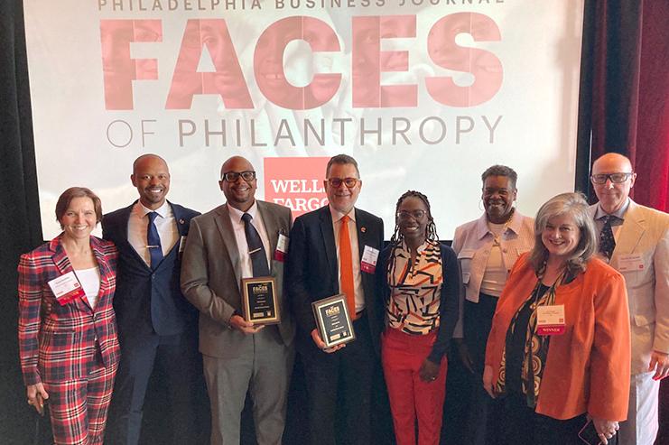 Group standing in front of a 'Faces of Philanthropy' banner. Two people are holding awards.