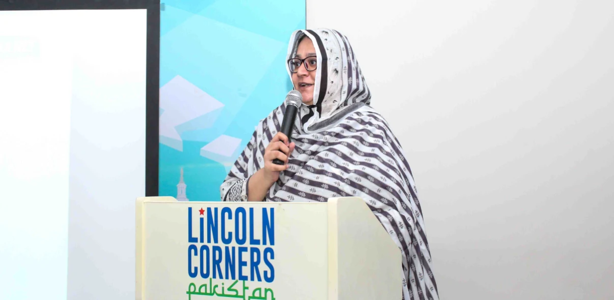 Faiza Yousuf standing at a podium, holding a microphone. Lincoln Corners Pakistan on the front