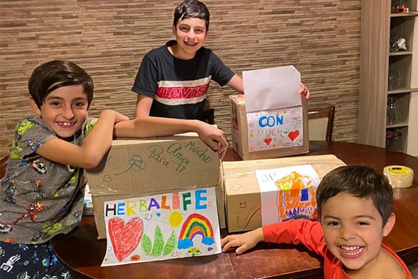 Three children at a table, boxes with Herbalife and drawings on them.