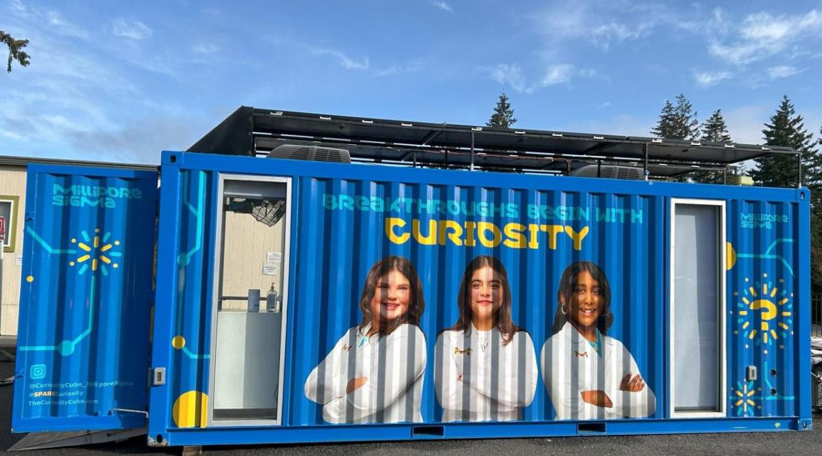 lays three The Curiosity Cube, a blue mobile science lab that displays three students in white lab coats and text that reads "Breakthroughs Begin With Curiosity"