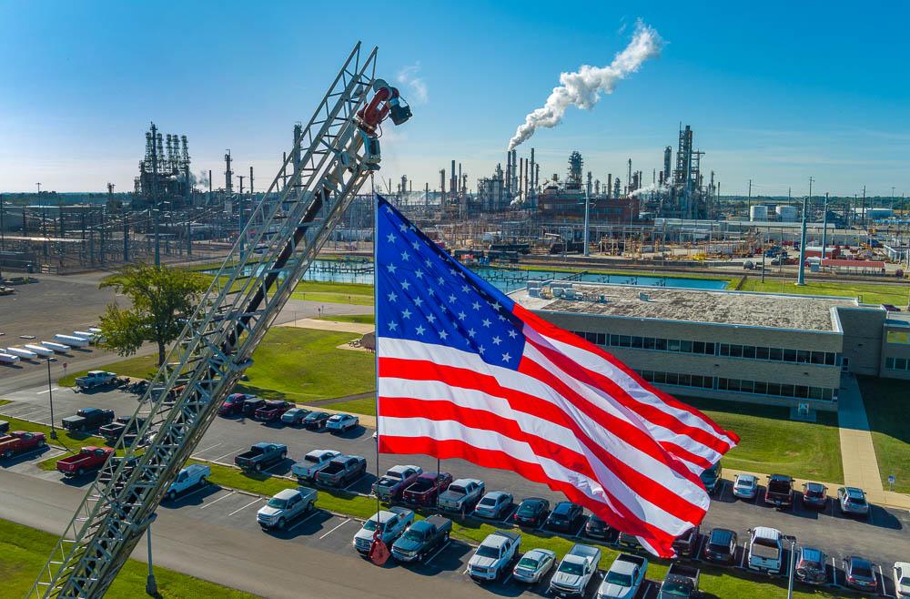 An American flag high above a parking lot with industrial plant behind it.