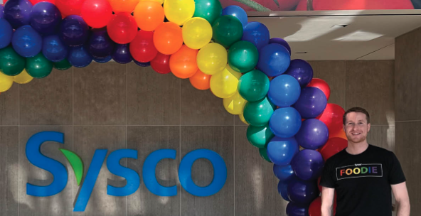 Person standing next to Sysco logo and rainbow of balloons. 