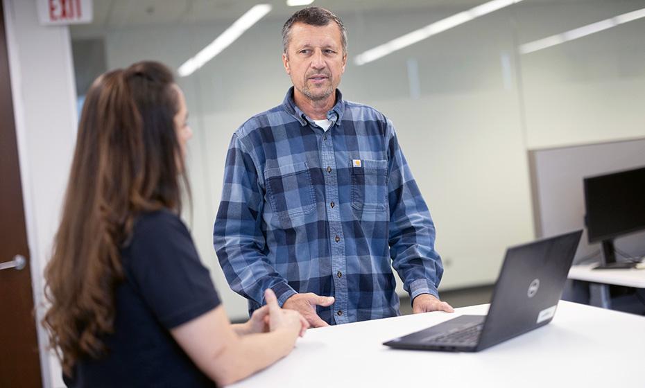 One person sat at a desk with a laptop, talking to a person who is stood beside them