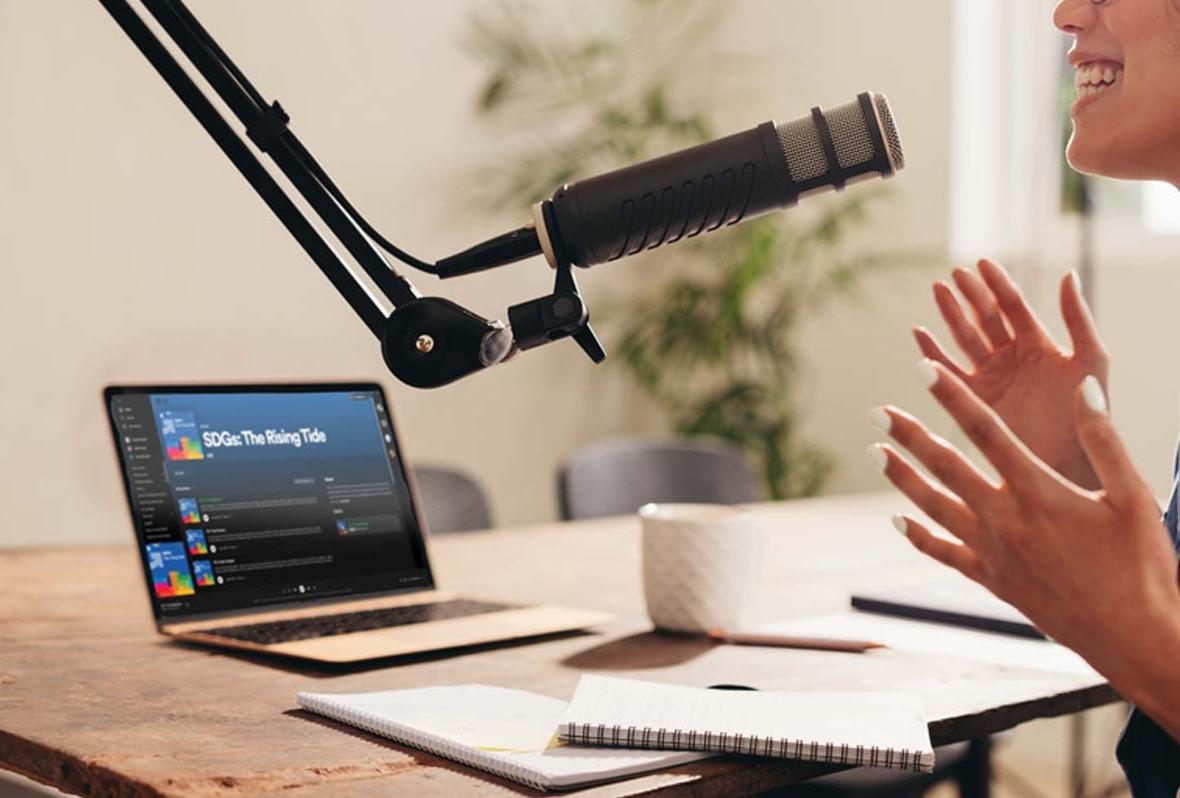 Catch up on 'The Rising Tide' - the podcast series on the SDGs, from Global Reporting Initiative person speaking into microphone with laptop next to them on a desk