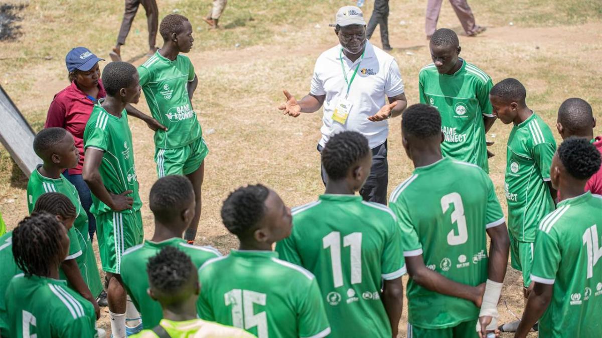 A soccer team huddled and the coach talking with them. They are wearing green uniforms with white writing, standing on a grass and sand field.