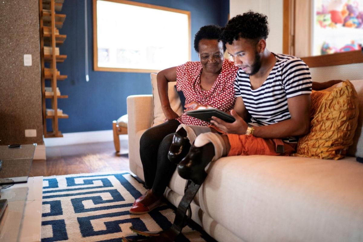 Photo of a woman sitting on a couch next to a man with prosthetic legs, both are looking at a tablet