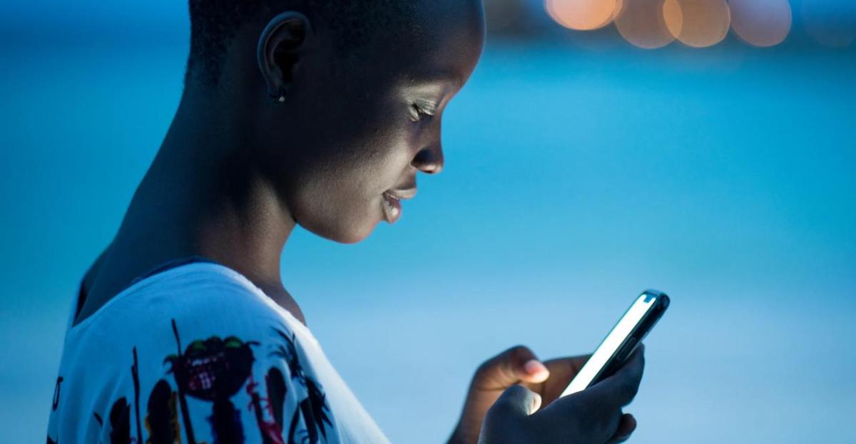 Close-up of a young woman using a smartphone at dusk, her face illuminated by the screen’s glow. She appears focused, with a soft blue background and blurred city lights in the distance.