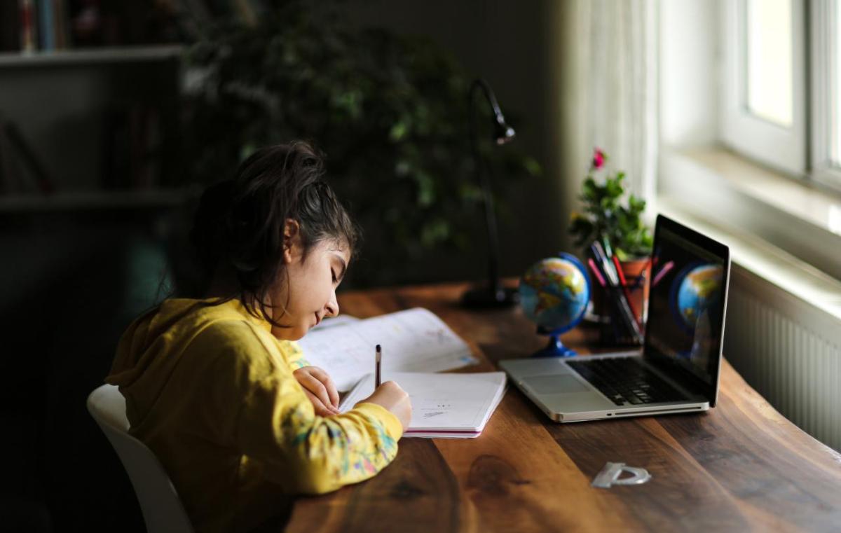 student writing on paper in front of a laptop