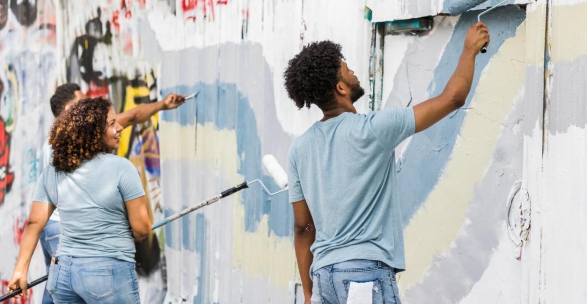 Group of young adults painting over a graffiti-covered wall with rollers and brushes, working together as part of a community mural or cleanup project. They wear casual clothes and appear engaged and cheerful.
