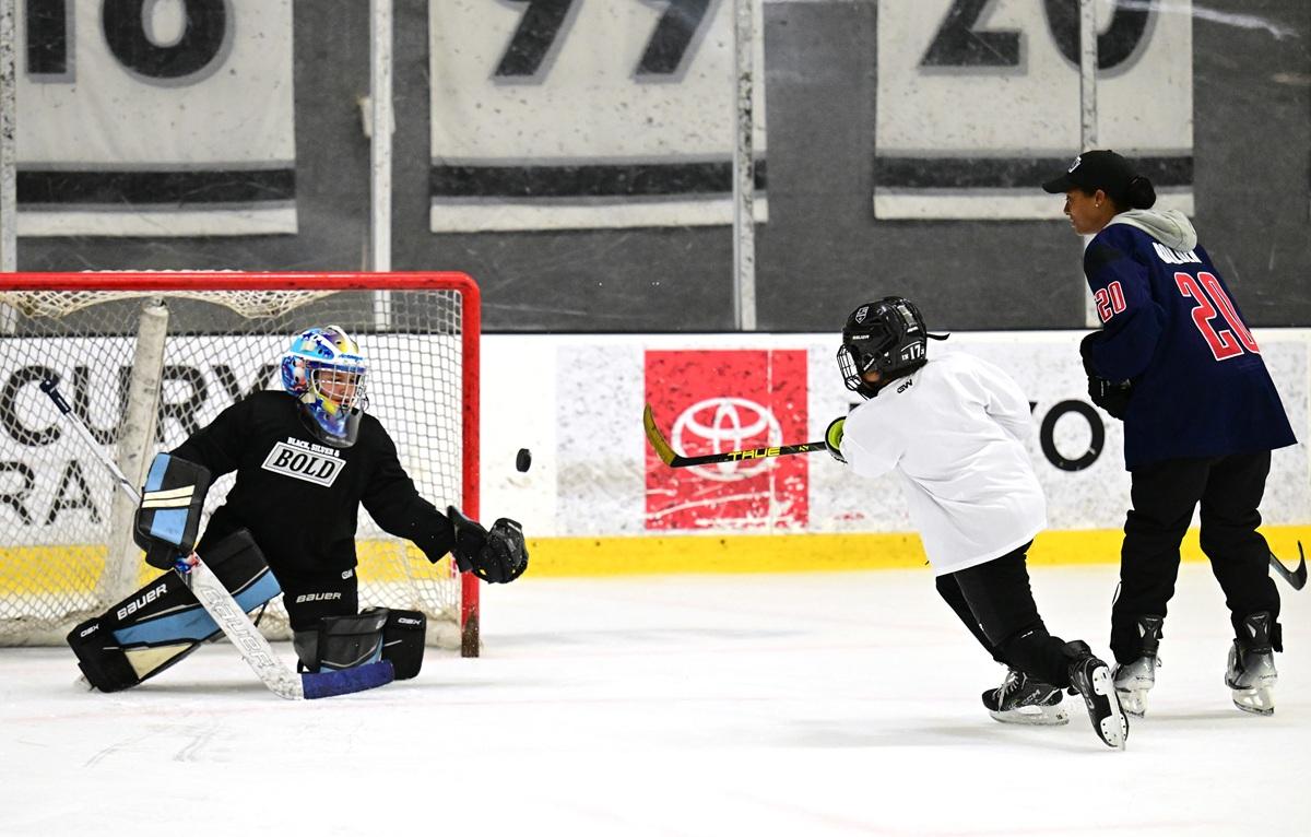 Youth hockey player takes a shot on goal as a goalie in Black, Silver & Bold gear makes a save, with a coach watching on the ice.