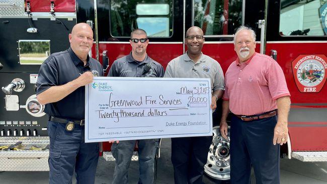 Four people standing in front of a fire truck, holding a large check.