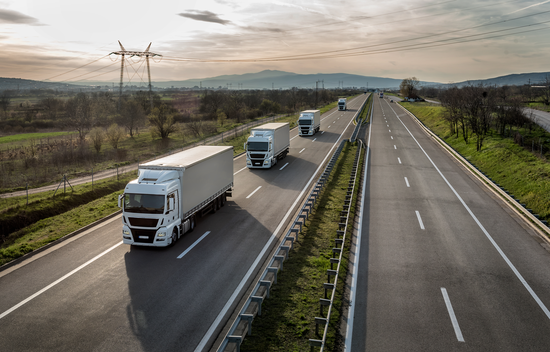 A row of semi trucks on a highway. Powerlines to the side and a scenic background.