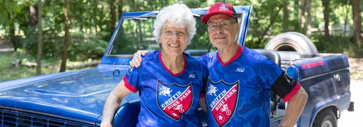 Gene and Donna Griffin, standing in front of a jeep, both wearing matching blue Griffin Bike Trail bike shirts