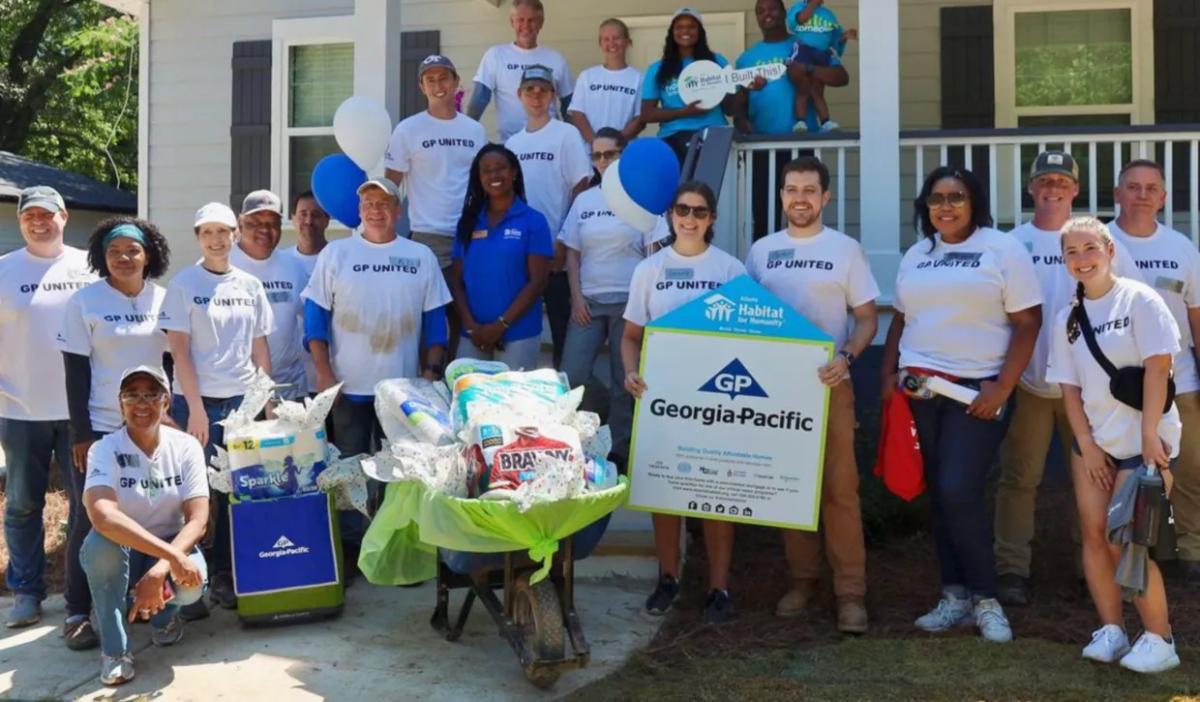 A group of volunteers in front of a house. All wearing shirts that say "GP United". A wheelbarrow full of supplies in front of them and a Georgia-Pacific sign being held by two.