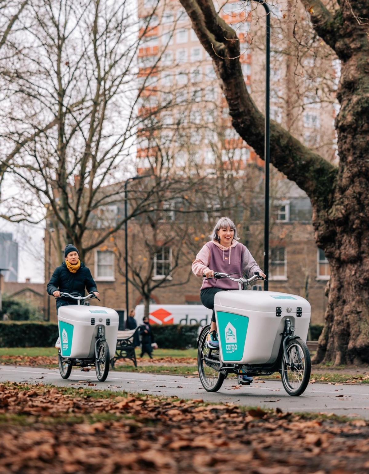 Two people riding cargo bikes through a park.