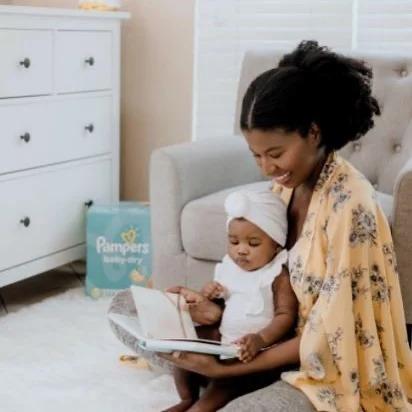 An adult and baby sitting on the floor reading a book. An upholstered chair and window behind them. A box of "Pampers" in the corner.