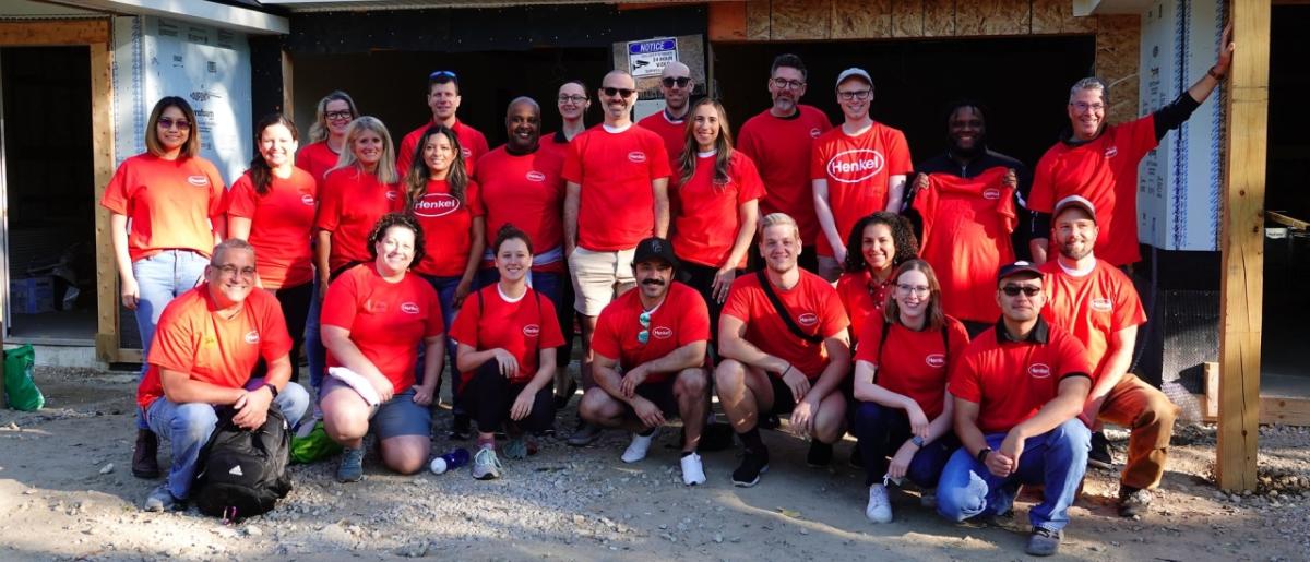 group of people outside a home being built, all are wearing matching orange shirts with Henkel logo