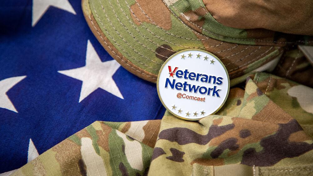 An american flag behind a camo patterned hat and shirt. A badge with "Veterans Network Comcast" on top.