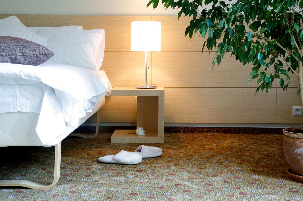 Horizontal view of a hotel room with a bed, side table, lamp, and slippers